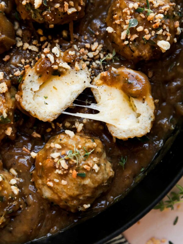 French onion meatballs in a large cast iron skillet that sits atop a light pink textured surface with a blue and white striped linen napkin placed underneath. The meatballs have been topped with toasty garlic herb breadcrumbs. One of the meatballs have been sliced in half and split open to reveal a melty gruyère cheese center.