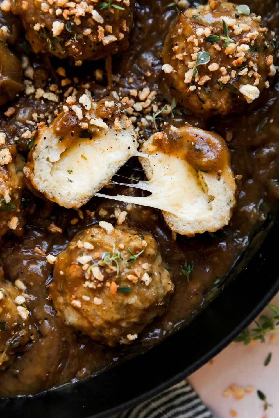 French onion meatballs in a large cast iron skillet that sits atop a light pink textured surface with a blue and white striped linen napkin placed underneath. The meatballs have been topped with toasty garlic herb breadcrumbs. One of the meatballs have been sliced in half and split open to reveal a melty gruyère cheese center.