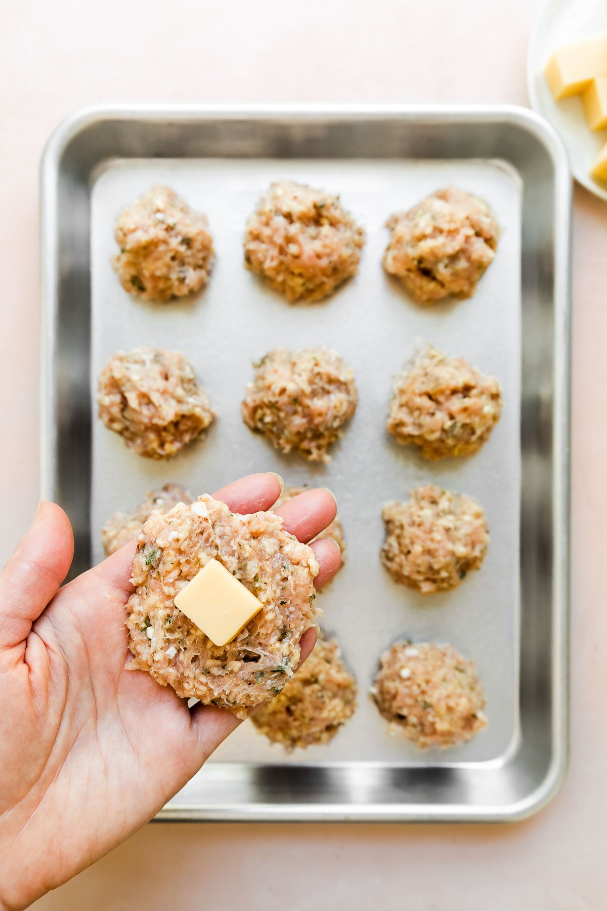 A woman's hand holds a small patty of chicken meatball mixture with a cube of gruyère cheese placed in the center above a pan of formed gruyère cheese stuffed chicken meatballs. The baking sheet sits atop a light pink surface with a small white plate of gruyère cheese cubes sitting alongside.