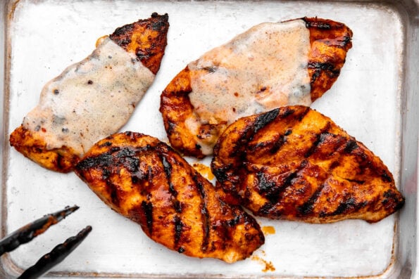 Four grilled buffalo chicken breasts arranged atop an aluminum baking sheet. Two of the chicken breasts are shown with melted cheese overtop. A pair of grilling tongs rests on the side of the baking sheet.
