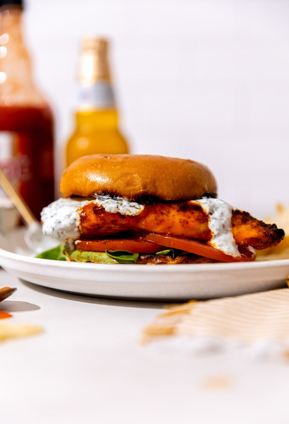 A finished buffalo chicken sandwich served on a white plate in between a toasted brioche bun with lettuce, tomato, and herb ranch yogurt drizzled on top. The white plate sits atop a creamy white textured surface with a small bowl of herb ranch yogurt, a bottle of Franks' RedHot Original, and a bottle of beer shown in the background.