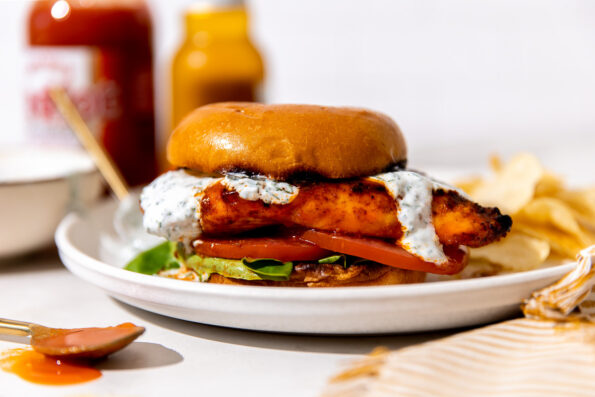 A finished buffalo chicken sandwich served on a white plate in between a toasted brioche bun with lettuce, tomato, and herb ranch yogurt drizzled on top and a side of potato chip. The white plate sits atop a creamy white textured surface with a white and gold linen napkin tucked underneath the plate. A small bowl of herb ranch yogurt, a bottle of Franks' RedHot Original, and a bottle of beer shown in the background while a gold spoonful of buffalo sauce rests atop the surface in the foreground.