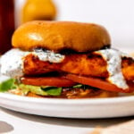 A finished buffalo chicken sandwich served on a white plate in between a toasted brioche bun with lettuce, tomato, and herb ranch yogurt drizzled on top and a side of potato chip. The white plate sits atop a creamy white textured surface with a white and gold linen napkin tucked underneath the plate. A small bowl of herb ranch yogurt, a bottle of Franks' RedHot Original, and a bottle of beer shown in the background while a gold spoonful of buffalo sauce rests atop the surface in the foreground.