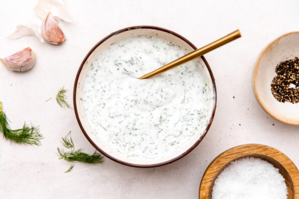 A small bowl filled with herby ranch yogurt sits atop a creamy white textured surface with a gold spoon resting inside the bowl for stirring. The bowl is surrounded by cloves of garlic, fresh dill, and two small pinch bowls filled with kosher salt and ground black pepper.