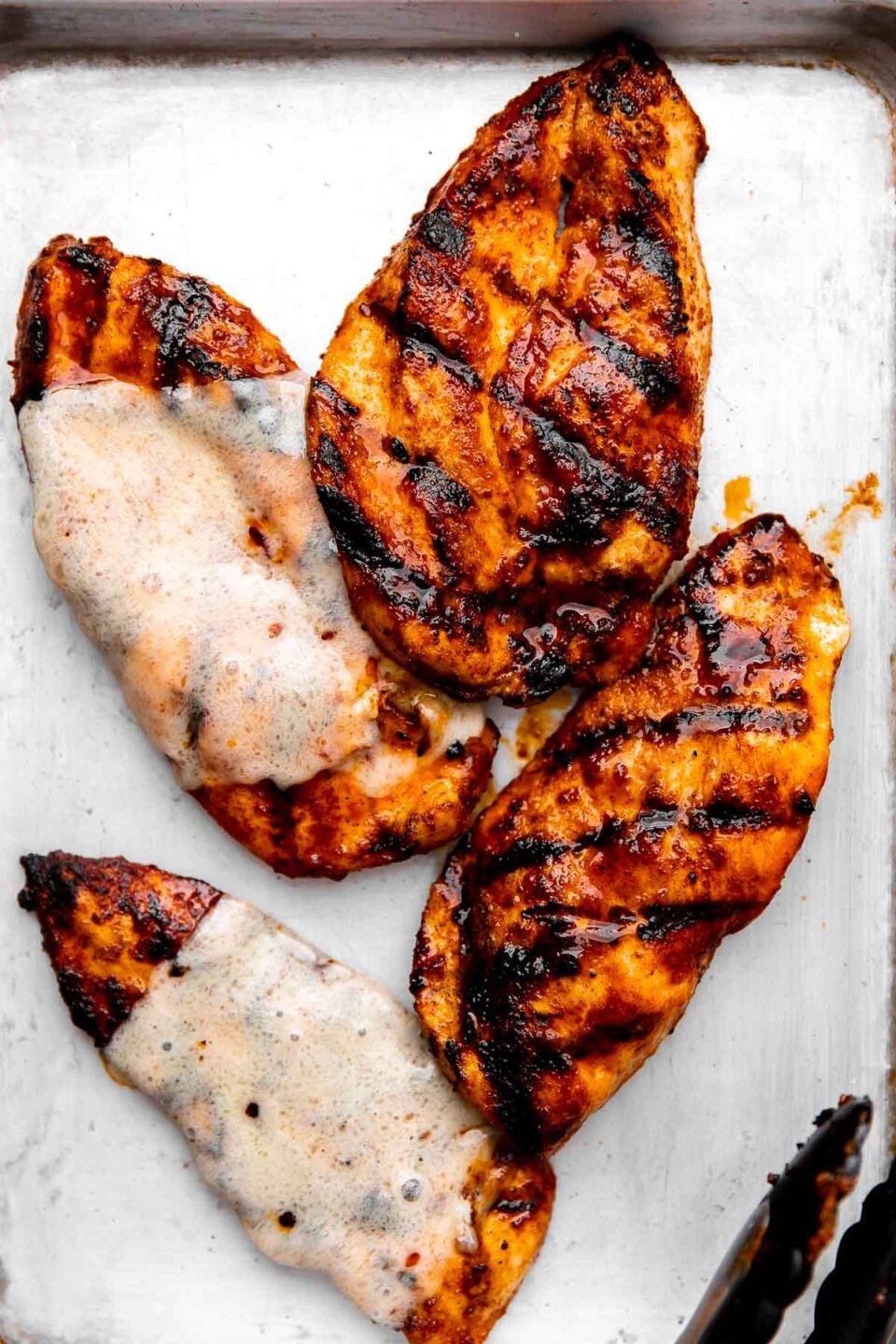 Four grilled buffalo chicken breasts arranged atop an aluminum baking sheet. Two of the chicken breasts are shown with melted cheese overtop. A pair of grilling tongs rests on the side of the baking sheet.