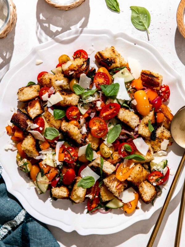 Grilled panzanella salad is served atop a white serving platter that sits atop a white textured surface. The panzanella salad has been garnished with fresh herbs and shaved asiago cheese. Two gold serving spoons rest atop the platter and the platter is surrounded by two drinking glasses, a small pinch bowl filled with kosher salt, and a blue linen napkin.