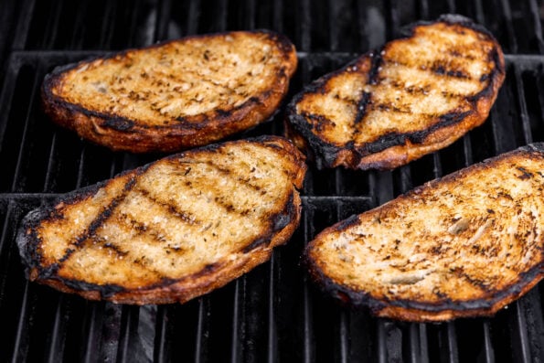 Four pieces of sliced sourdough bread are being grilled atop gas grill grates for grilled bread salad.