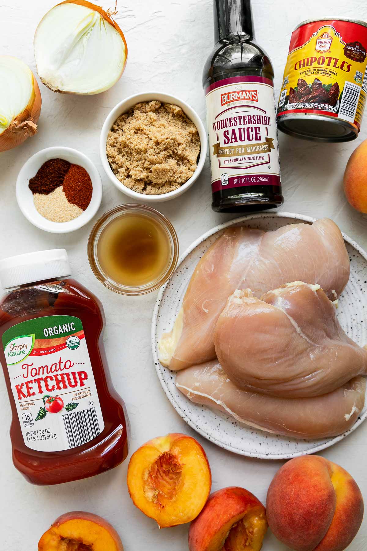 Peach bbq chicken and peach bbq sauce ingredients arranged on a creamy white textured surface: bonless, skinless chicken breasts, Stonemill Chili Powder, Stonemill Garlic Powder, Stonemill Paprika, Simply Nature Avocado Oil, peaches, Pueblo Lindo Chipotle Peppers, brown sugar, Simply Nature Organic Apple Cider Vinegar, Simply Nature Organic Ketchup, Burman's Worcestershire Sauce, kosher salt, and ground black pepper.