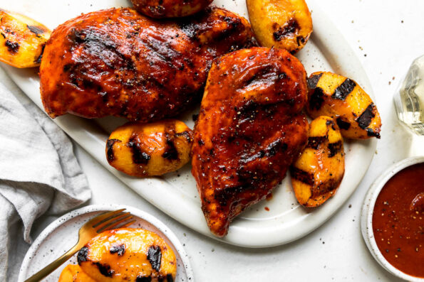 Three grilled peach bbq chicken breasts are arranged atop a white serving platter. The platter sits atop a creamy white textured surface surrounded by a glass of white wine, a small white ceramic plate with grilled peaches a gold fork resting atop, a small white ceramic bowl filled with chipotle peach bbq sauce, a glass of white wine, and a gray linen napkin.