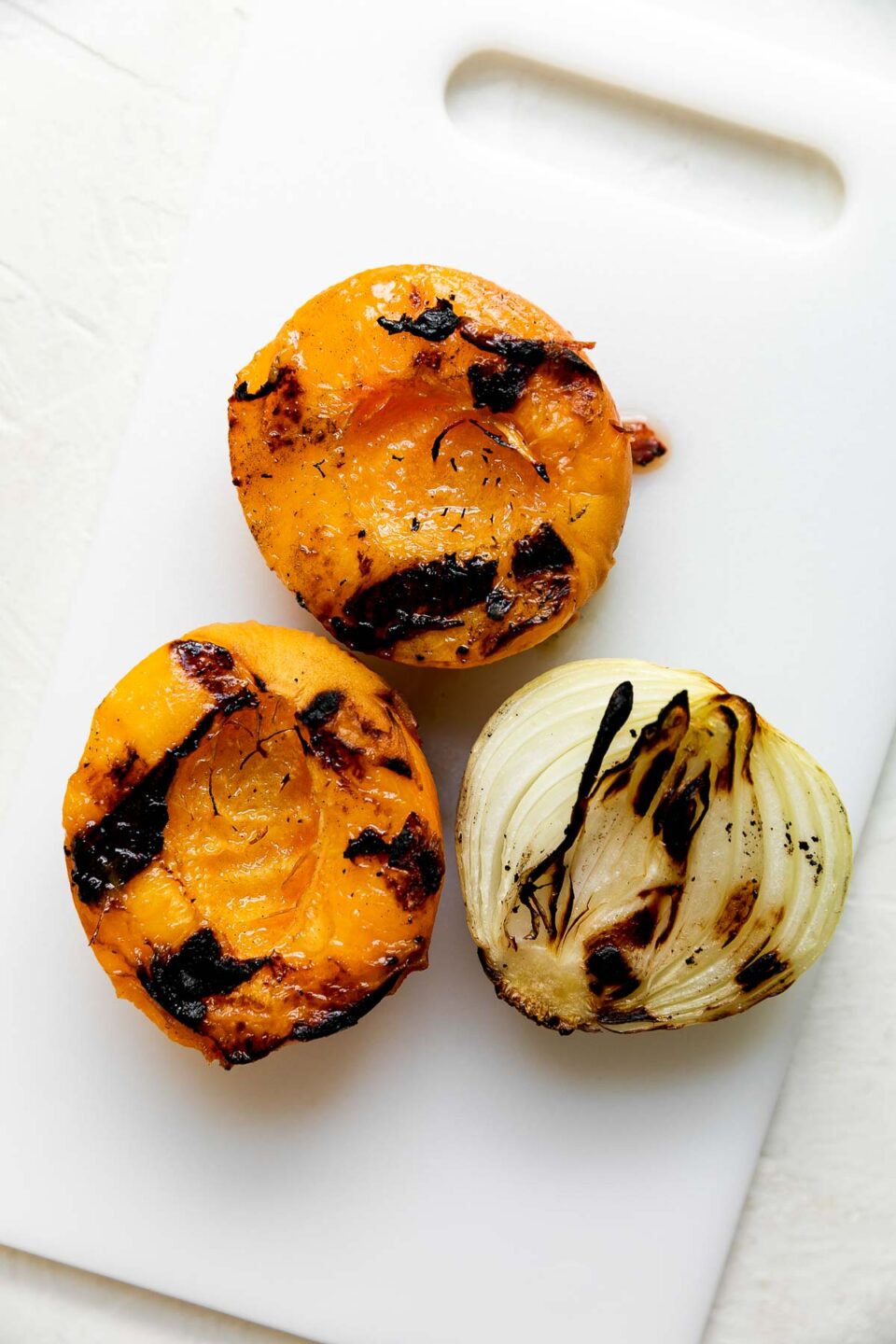 Two grilled peach halves and one half of a grilled onion sit atop a white plastic cutting board that sits atop a creamy white textured surface.