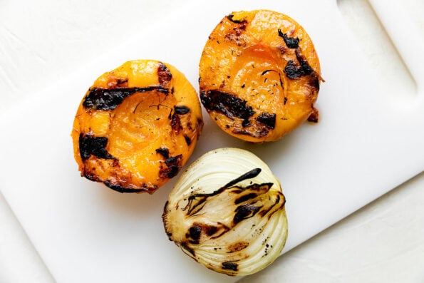Two grilled peach halves and one half of a grilled onion sit atop a white plastic cutting board that sits atop a creamy white textured surface.