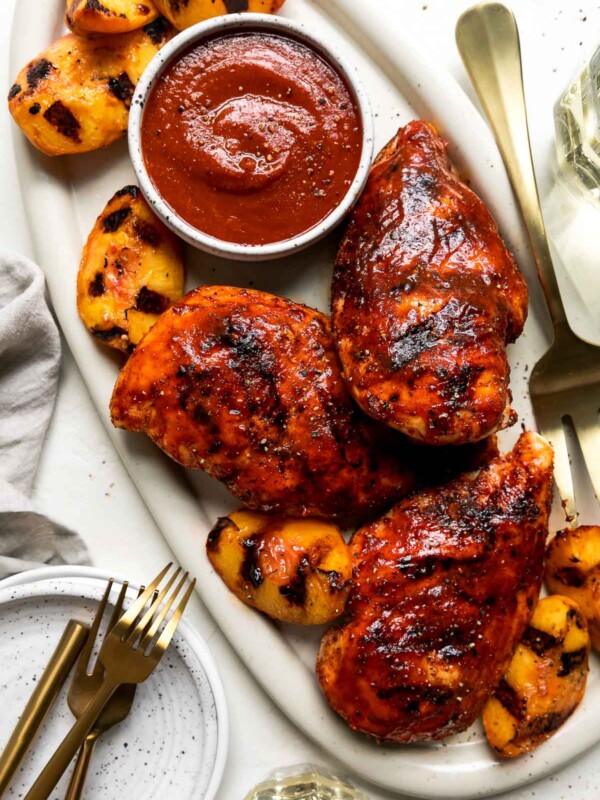 Three grilled peach bbq chicken breasts are arranged atop a white serving platter with grilled peach halves and a small white ceramic bowl filled with chipotle peach BBQ sauce. The platter sits atop a creamy white textured surface with a gold serving fork resting atop the platter. The serving platter & peach chicken are surrounded by two glasses of white wine, gold silverware atop a small stack of ceramic plates, and a gray linen napkin.