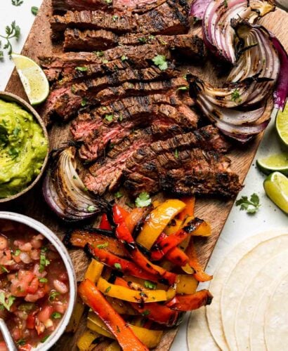 Grilled Steak fajitas cut into thin strips are served on a rectangular wood platter surrounded by grilled fajita veggies: bell peppers and onions. Fresh lime wedges surround the platter while a small wooden pinch bowl filled with guacamole and a small white ceramic bowl filled with fresh salsa rests atop the platter next to the steak and veggies. Fresh cilantro, a pile of corn tortillas and a small white ceramic pinch bowl filled with cotija cheese surround the platter at center.