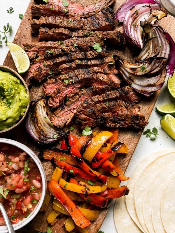 Grilled Steak fajitas cut into thin strips are served on a rectangular wood platter surrounded by grilled fajita veggies: bell peppers and onions. Fresh lime wedges surround the platter while a small wooden pinch bowl filled with guacamole and a small white ceramic bowl filled with fresh salsa rests atop the platter next to the steak and veggies. Fresh cilantro, a pile of corn tortillas and a small white ceramic pinch bowl filled with cotija cheese surround the platter at center.
