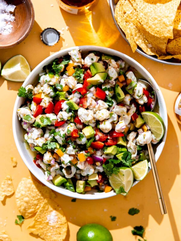A side angle shot of finished shrimp ceviche with avocado fills a large white ceramic serving bowl. A gold serving spoon rests inside the bowl and the shrimp ceviche has been garnished with slices of fresh lime and cilantro. The bowl sits atop a yellow surface surrounded by yellow corn tortilla chips in a bowl, a small wooden pinch bowl filled with kosher salt, a beer bottle and a beer bottle cap, and lime wedges.