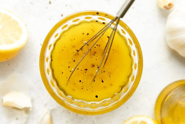 White balsamic vinaigrette fills a small clear glass bowl with a small metal whisk resting inside. The bowl sits atop a creamy white textured surface surrounded by lemon halves, loose garlic cloves, and a bottle of olive oil.