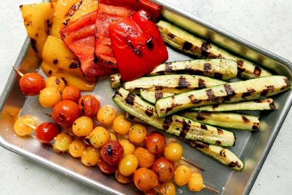 Grilled skewers of cherry tomatoes, grilled pieces of yellow and red bell peppers, and grilled zucchini spears are arranged atop a small alumninum baking sheet. The baking sheet sits atop a creamy white textured surface.