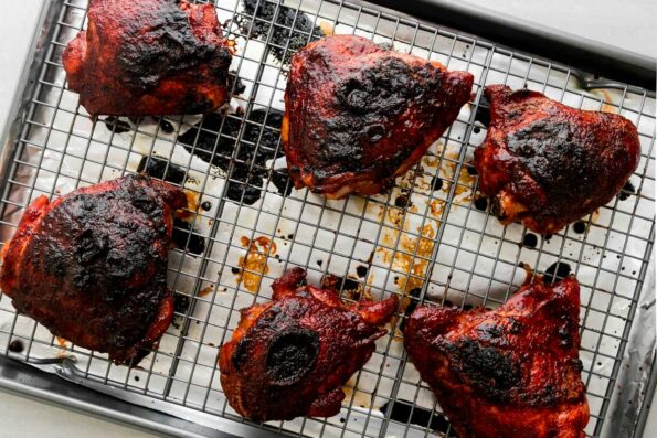 Six pieces of char siu chicken that have been roasted, broiled, and basted with char siu sauce sit atop a wire rack that sits above an aluminum lined baking sheet. The baking sheet sits atop a creamy white textured surface.