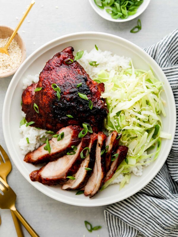 Char siu chicken served atop rice and thinly sliced and sauteed cabbage on a white ceramic plate. The plate sits atop a creamy white textured surface surrounded by a blue and white linen napkin, a small white ceramic bowl filled with thinly sliced green onion, a small wooden pinch bowl filled with sesame seeds with a small gold spoon resting inside, and two gold forks surround the bowl at center.