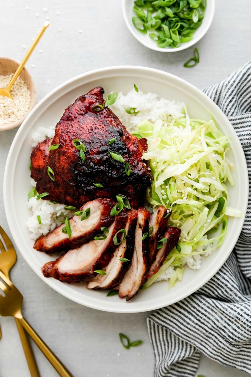 Char siu chicken served atop rice and thinly sliced and sauteed cabbage on a white ceramic plate. The plate sits atop a creamy white textured surface surrounded by a blue and white linen napkin, a small white ceramic bowl filled with thinly sliced green onion, a small wooden pinch bowl filled with sesame seeds with a small gold spoon resting inside, and two gold forks surround the bowl at center.