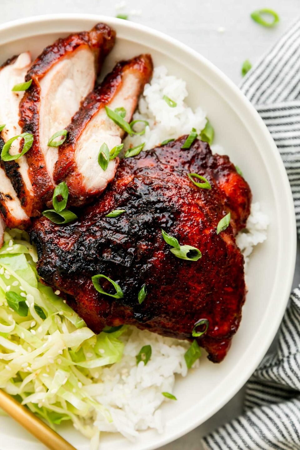 An overhead and close up shot of char siu chicken served atop rice and thinly sliced and sauteed cabbage on a white ceramic plate. The plate sits atop a creamy white textured surface surrounded by a blue and white linen napkin.