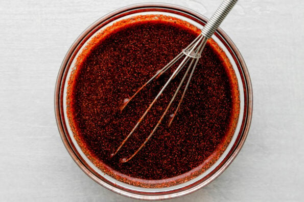 Char siu marinade mixed in a small clear glass mixing bowl sits atop a creamy white textured surface. A small metal whisk rests inside of the bowl for stirring.