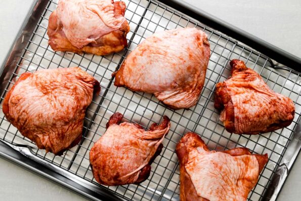 Bone-in, skin-on chicken thighs that have been marinated in char siu marinade sit atop a rack that sits above an aluminum lined baking sheet. The baking sheet sits atop a creamy white textured surface.