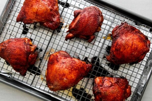 Six pieces of roasted char siu chicken sit atop a wire rack that sits above an aluminum lined baking sheet. The baking sheet sits atop a creamy white textured surface.