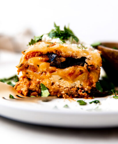 A single piece of lasagna with eggplant sits atop a white ceramic plate. A small wooden bowl filled with a mixed greens salad sits atop the plate and a gold spoon rests atop the plate alongside the lasagna. The plate sits atop a creamy white textured surface with a plaid linen napkin in the background rests out of focus.