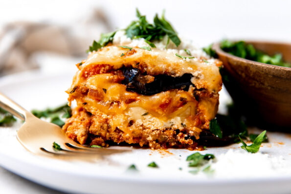 A single piece of eggplant lasagna sits atop a white ceramic plate. A small wooden bowl filled with a mixed greens salad sits atop the plate and a gold spoon rests atop the plate. The plate sits atop a creamy white textured surface with a plaid linen napkin in the background rests out of focus.
