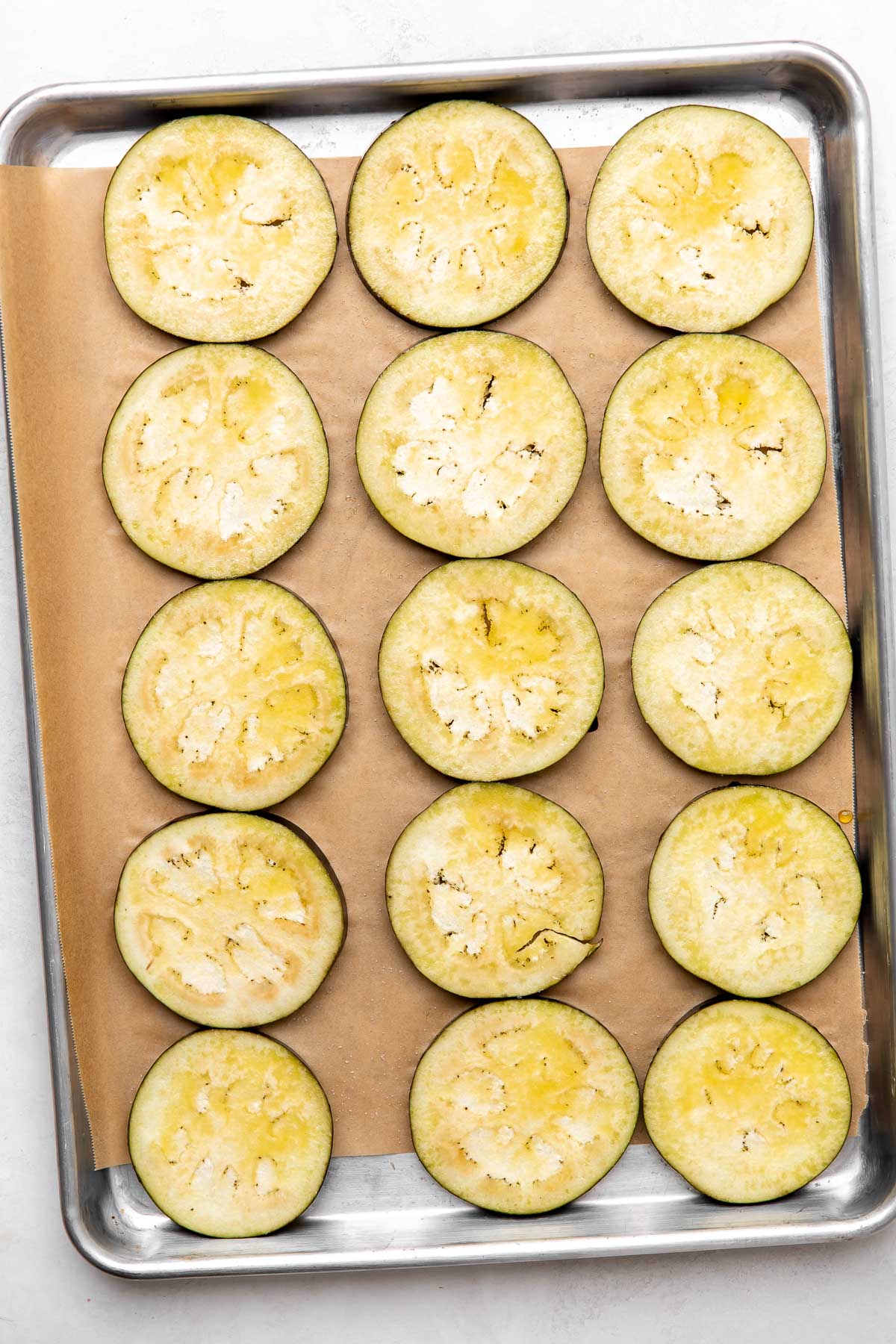 Eggplant rounds arranged on a parchment lined baking sheet sits atop a creamy white textured surface.