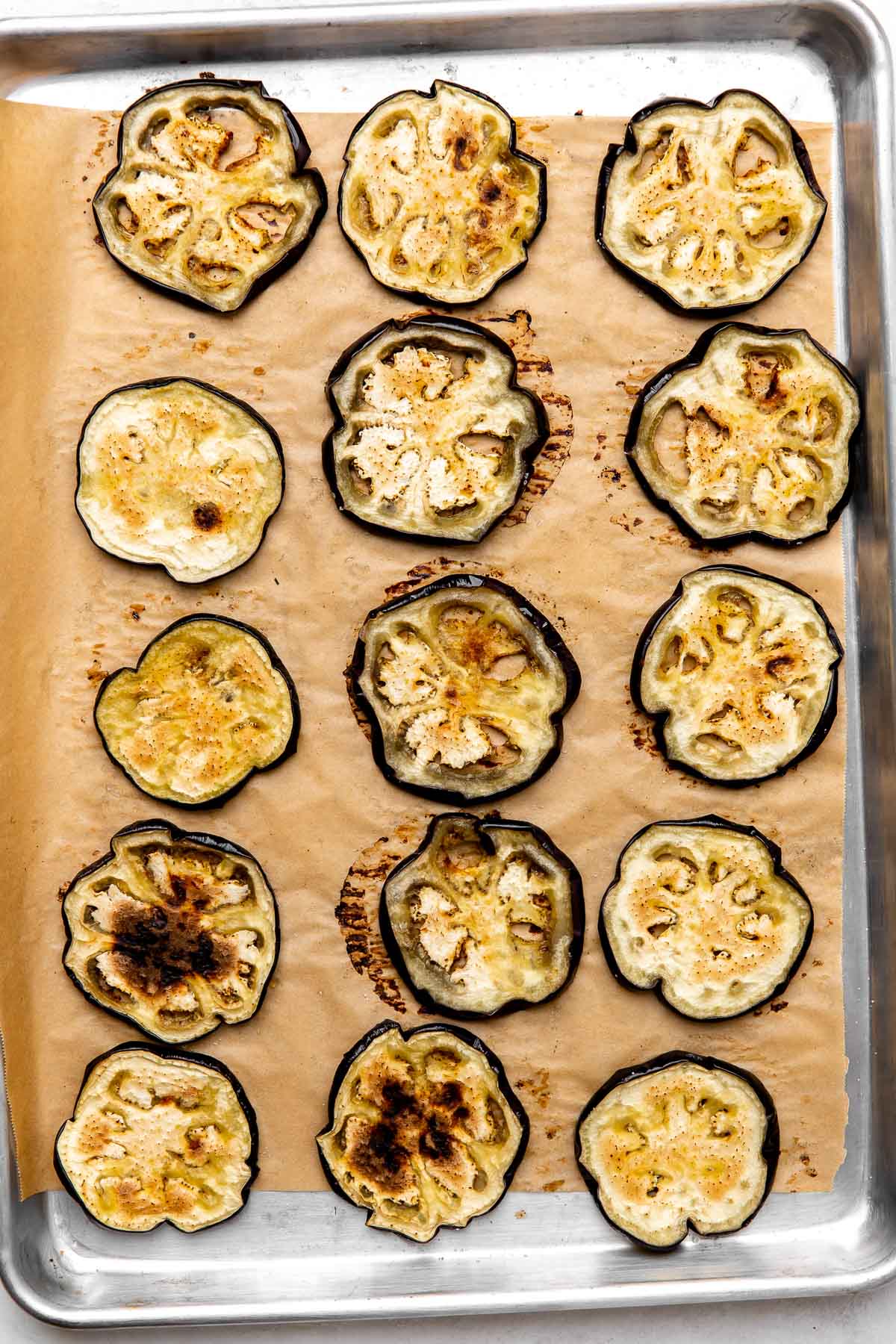 Sliced roasted eggplant rounds arranged on a parchment lined baking sheet. The baking sheet sits atop a creamy white textured surface.