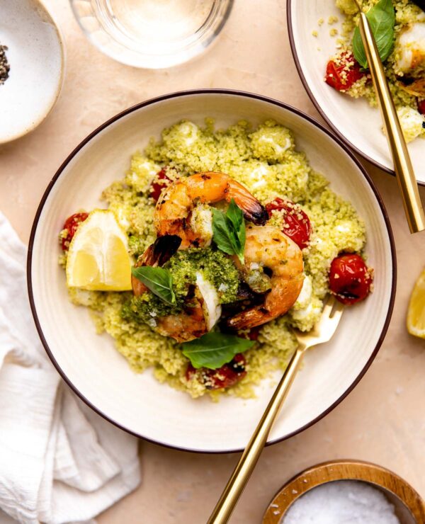 An overhead shot of grilled shrimp with pesto rests atop a pile of Caprese couscous with burst grilled cherry tomatoes and mozzarella pearls inside of a small cream colored bowl. The shrimp is garnished with small fresh basil leaves and a few lemon wedges and a gold fork rests inside of the bowl. A cream colored linen napkin, a drinking glass, two small pinch bowls filled with salt and pepper, and another serving of pesto shrimp surround the bowl at center.