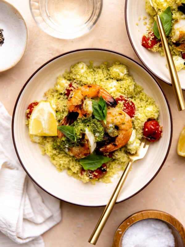 An overhead shot of grilled shrimp with pesto rests atop a pile of Caprese couscous with burst grilled cherry tomatoes and mozzarella pearls inside of a small cream colored bowl. The shrimp is garnished with small fresh basil leaves and a few lemon wedges and a gold fork rests inside of the bowl. A cream colored linen napkin, a drinking glass, two small pinch bowls filled with salt and pepper, and another serving of pesto shrimp surround the bowl at center.