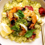 A close up, overhead, and macro shot of grilled shrimp with pesto atop a pile of Caprese couscous with burst grilled cherry tomatoes and mozzarella pearls fills a cream colored bowl. The shrimp is garnished with small fresh basil leaves and a few lemon wedges and a gold fork rests inside of the bowl. A cream colored linen napkin, a small pinch bowl, and another bowl filled with another serving of grilled pesto shrimp surrounds the bowl at center.