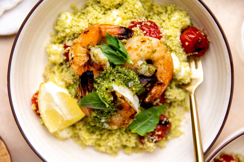 A close up, overhead, and macro shot of grilled shrimp with pesto atop a pile of Caprese couscous with burst grilled cherry tomatoes and mozzarella pearls fills a cream colored bowl. The shrimp is garnished with small fresh basil leaves and a few lemon wedges and a gold fork rests inside of the bowl. A cream colored linen napkin, a small pinch bowl, and another bowl filled with another serving of grilled pesto shrimp surrounds the bowl at center.