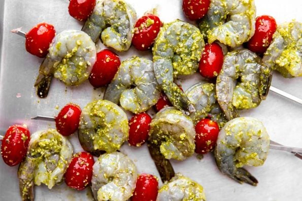 A close up shot of pesto shrimp and cherry tomatoes threaded on a stainless steel skewers rest atop an aluminum baking sheet.