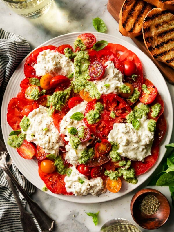 Heirloom tomato burrata salad arranged on large white serving platter sits atop a white and gray marbles surface. The platter is surrounded by a wooden serving board topped with grilled bread, fresh basil, a small wooden pinch bowl filled with ground black pepper, two small drinking glasses filled with white wine, a gray and white striped linen napkin, and silver servingware.