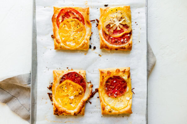 Four baked heirloom tomato tarts are arranged atop a parchment lined baking sheet. The baking sheet sits atop a creamy white textured surface with a gray linen napkin placed underneath.