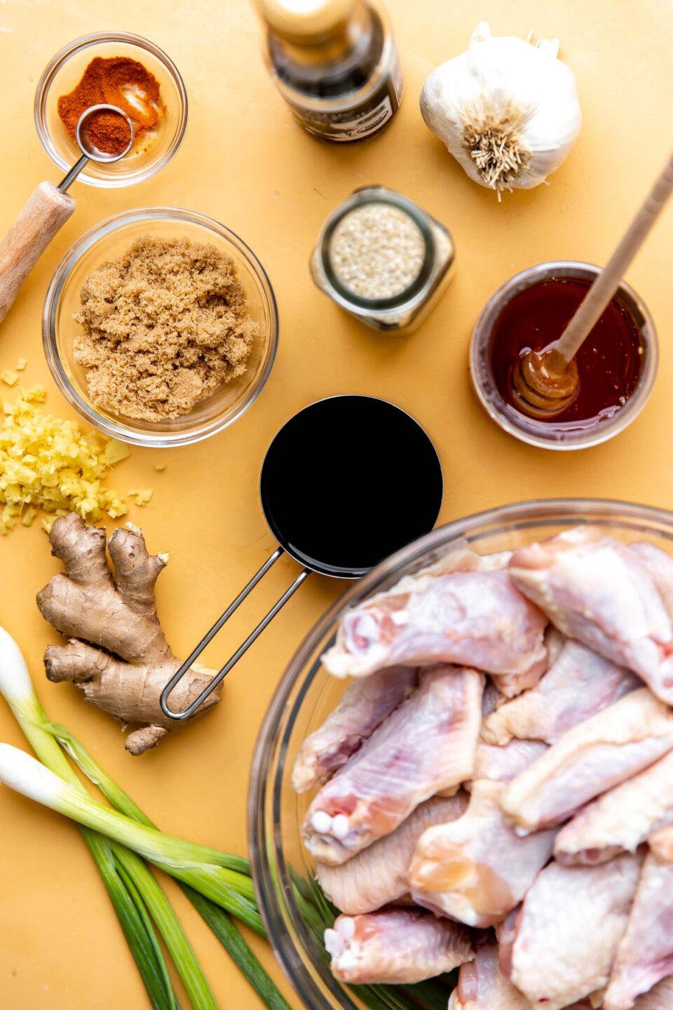 Honey sesame chicken wings ingredients arranged on a yellow textured surface: chicken wing sections, soy sauce, honey, dark brown sugar, sesame oil, fresh ginger, cayenne pepper, garlic, sesame seeds, and green onions.