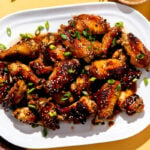 Honey sesame chicken wings served atop a white serving platter and garnished with additional sesame seeds and thinly sliced green onion. The platter sits atop a textured yellow surface surrounded by amber drinking glasses, a red and white plate with wooden chopsticks, a beer bottle cap, and a red and white linen napkin.