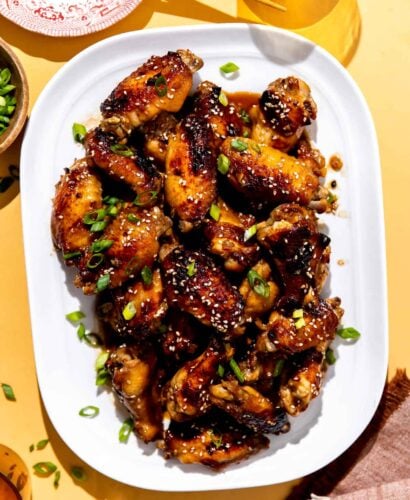 Honey sesame chicken wings served atop a white serving platter and garnished with additional sesame seeds and thinly sliced green onion. The platter sits atop a textured yellow surface surrounded by amber drinking glasses, a red and white plate with wooden chopsticks, a beer bottle cap, and a red and white linen napkin.