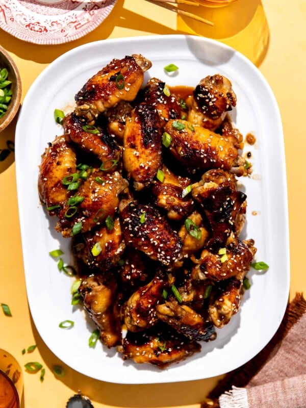 Honey sesame chicken wings served atop a white serving platter and garnished with additional sesame seeds and thinly sliced green onion. The platter sits atop a textured yellow surface surrounded by amber drinking glasses, a red and white plate with wooden chopsticks, a beer bottle cap, and a red and white linen napkin.