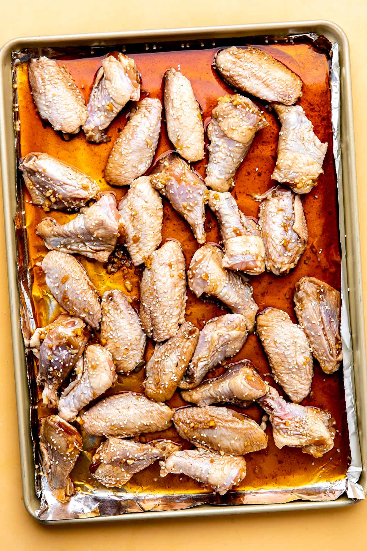 How to make baked sesame wings, step 2: Prep the chicken wings for baking. Chicken wings sections that have been marinated in honey sesame marinade are spread out across an aluminum foil lined baking sheet in a single layer. The baking sheet sits atop a yellow textured surface.