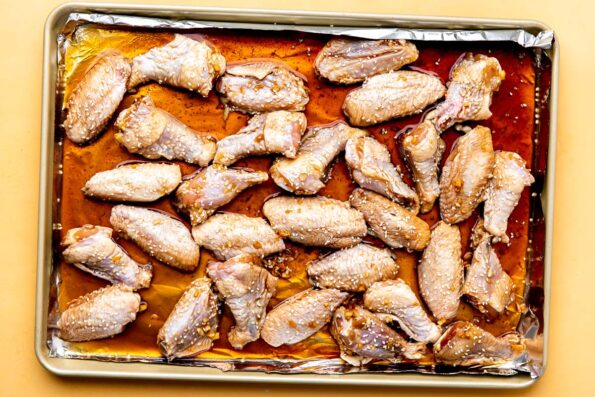 How to make baked sesame wings, step 2: Prep the chicken wings for baking. Chicken wings sections that have been marinated in honey sesame marinade are spread out across an aluminum foil lined baking sheet in a single layer. The baking sheet sits atop a yellow textured surface.