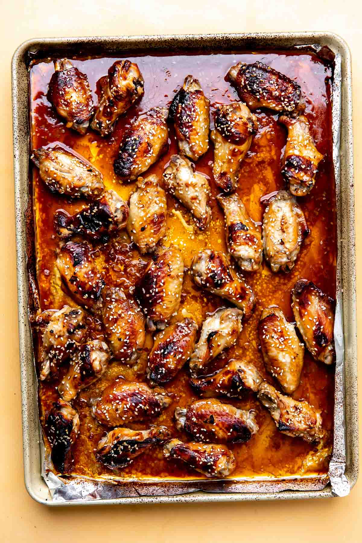 Baked honey sesame chicken wings are spread across an aluminum foil lined baking sheet in a single layer. The baking sheet sits atop a yellow textured surface.