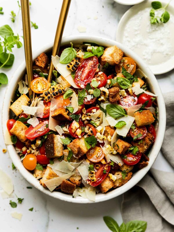 A finished cherry tomato salad fills a large white serving bowl atop a white and gray marble surface. Two gold spoons rest inside of the bowl and a light gray linen napkin sits alongside. Loose fresh herbs and two small white pinch bowls filled with kosher salt and ground black pepper surround the bowl at center.