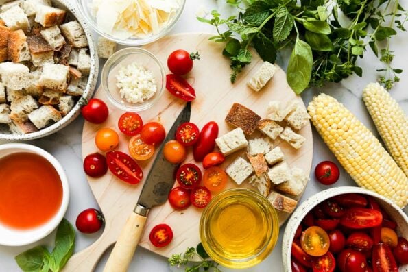 Summer tomato salad ingredients arranged on a white and gray marble surface with a wooden cutting board resting on top: cherry tomatoes, garlic, olive oil, red wine vinegar, sourdough bread, sweet corn, fresh herbs, asiago or parmesan cheese.