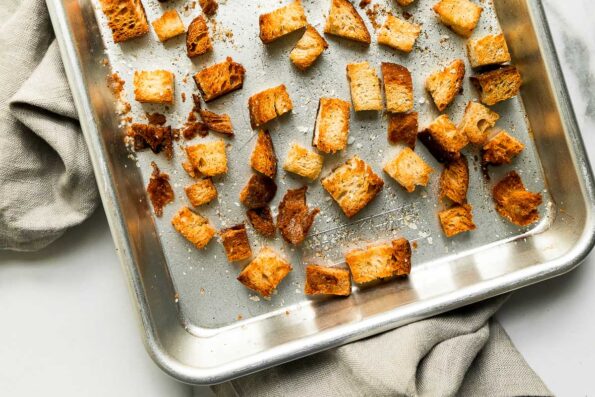 How to make a cherry tomato salad, step 3: Toast the bread cubes. Small pieces of toasted sourdough bread are arranged atop a small aluminum baking sheet. The baking sheet sits atop a white and gray marble surface with a light gray linen napkin placed underneath the sheet pan.