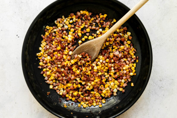 Pancetta sweet corn topping for sweet corn risotto sits inside of a gold non-stick skillet. The skillet sits atop a creamy white textured surface with a wooden spoon resting inside of the skillet for stirring.
